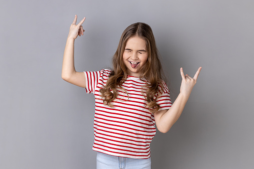 Portrait of little girl wearing striped T-shirt showing rock and roll gesture heavy metal sign, enjoys favorite music, has fun, exclaims from joy. Indoor studio shot isolated on gray background.