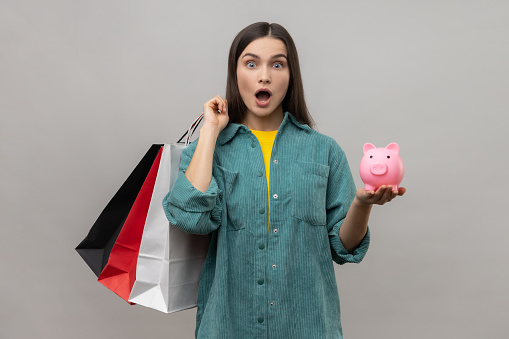 Amazed astonished woman with dark hair holding shopping bags and piggy bank, cashback from buying purchases, wearing casual style jacket. Indoor studio shot isolated on gray background.