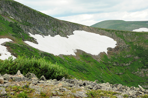 Remains of snow on the slope of a gentle hill overgrown with grass and bushes under a cloudy summer sky. Ivanovskie lakes, Khakassia, Siberia, Russia.