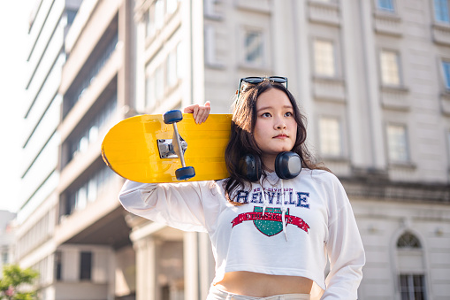 Young woman holding a skateboard on shoulder, having fashion glasses and headphones looking away