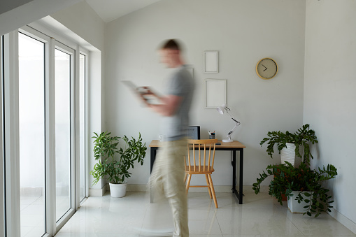 Blurred motion of businessman with tablet computer walking to big apartment window