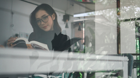 Beautiful asian girl reading a book shot over glass wall with surrounding reflections in a cafe