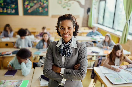 Happy African American female teacher standing on a class in front of her elementary students and looking at camera.