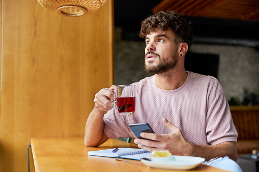 Young pensive man drinking tea and using mobile phone in a café. Copy space.