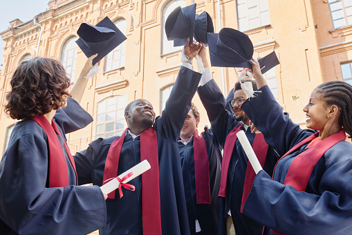 Multiethnic group of students in robes raising their hats up and celebrating their graduation