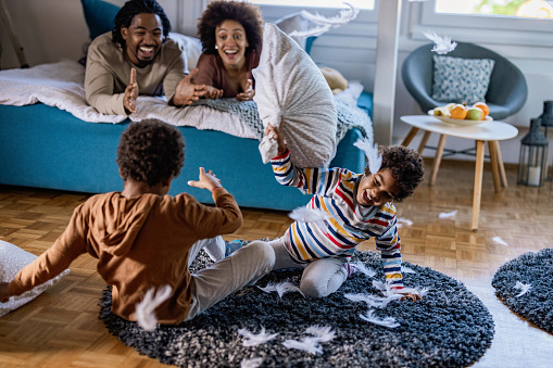Carefree African American kids having fun during a pillow fight at home. Their parents are in the background.