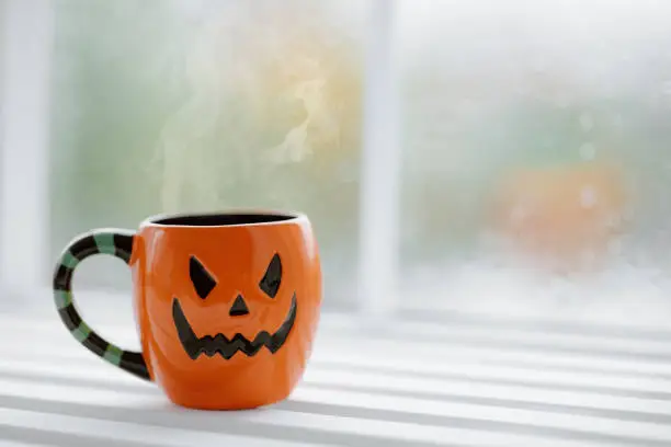 Photo of Close up of cup in shape of pumpkin Jack lantern with hot coffee on windowsill
