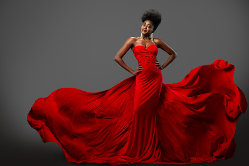 Fashion Woman in Red flying Dress with Afro Hairstyle. Dark Skinned Model in Silk Long Gown over Gray background. Happy elegant Lady wearing Luxury Clothes