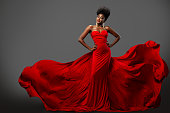 Fashion Woman in Red flying Dress with Afro Hairstyle. Dark Skinned Model in Silk Long Gown over Gray background. Happy elegant Lady wearing Luxury Clothes