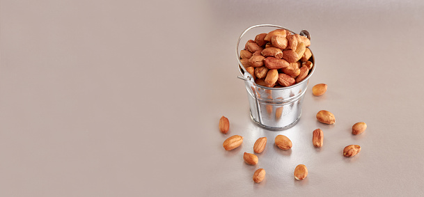 A small metal bucket filled with raw peanuts on a colored background.