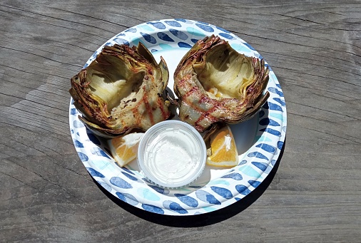 Grilled globe artichokes with sipping sauce and orange wedges on paper plate