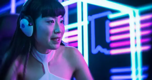 Photo of Portrait of a Beautiful Japanese Young Female Wearing Headphones, Talking with Friends Online on a Computer. Popular Asian Girl Streaming From a Futuristic Neon Room. Gamer Playing Video Games