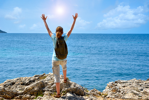 Woman with backpack and hands up standing on the rock near the sea and enjoying the beautiful view. Back view. Travel and active lifestyle concept.