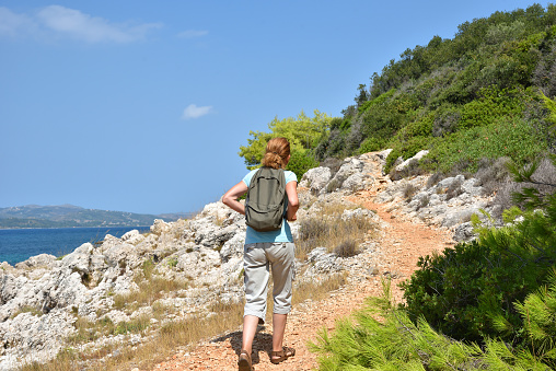 Woman with backpack walking on the coastal path near the sea and enjoying the nature. Back view. Travel and active lifestyle concept.