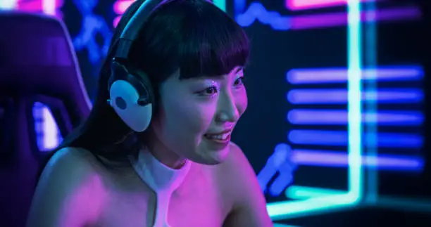 Photo of Portrait of a Beautiful Japanese Young Female Wearing Headphones, Talking with Friends Online on a Computer. Popular Asian Girl Streaming From a Futuristic Neon Room. Gamer Playing Video Games