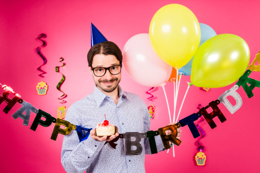 Happy birthday! Geek man with horn rimmed glasses, blue party hat, small Birthday Cake with cherry and five party multicolored decoration balloons in hand, isolated on pink background.