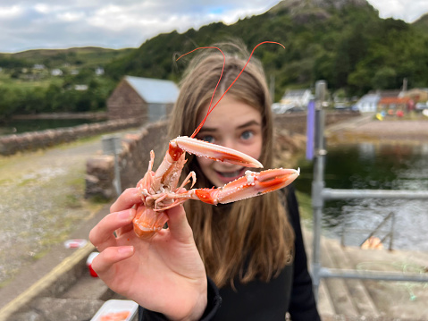 A young girl is showing a cooked langoustine close up to the camera. She is looking as if she is trying to scare the photographer. In the background is Lower Diabeg in the Scottish highlands.
