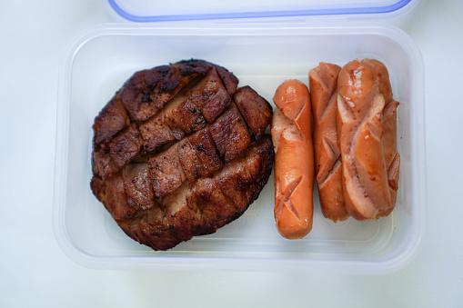 Top view of a homemade lunchbox containing grilled pork bacon and smoked pork sausage.