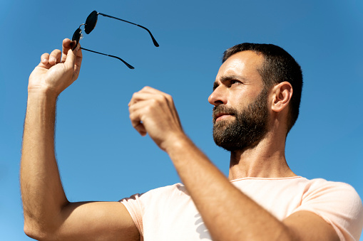 Portrait of handsome bearded man holding stylish sunglasses against blue sky