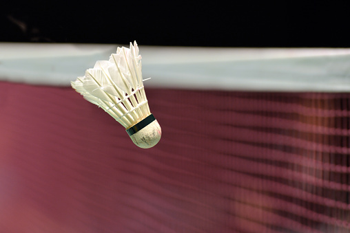 Closeup of a white shuttlecock for playing badminton in front of the purple netting on an indoor badminton court