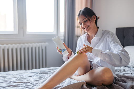 Beautiful young woman applying a moisturizer on her legs while sitting on the bed in her bedroom.