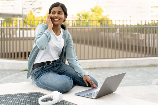 istock Smiling Indian woman using laptop computer, talking on mobile phone working project 1675819958