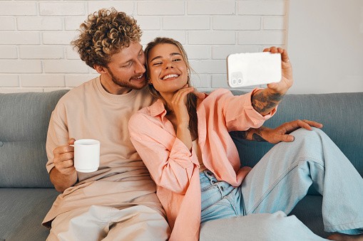 A young couple in love sitting on the living room sofa in a cozy home and hugging, romantically spending time together, drinking morning coffee and taking a selfie.