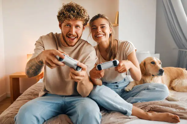 Young couple in love playing video games while sitting on the sofa in the bedroom, having fun and enjoying each other on their weekends.