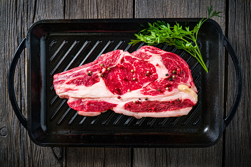 Beef rib steak raw vivid on a cast iron grill black background uncooked with parsley herbs on rustic dark wood table board background, top view flatlay