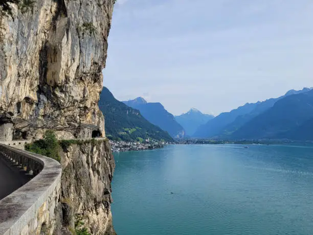 Clear, bright blue Swiss lake on a hot summer's day with a balcony road and tunnel in the foreground. In the distance are the Swiss Alps, highlighted against the summer sunshine.