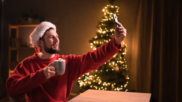 istock Happy young man with cup of cocoa holding video call conversation using cellphone apps, recording streaming online video in social network or does self-portrait on Christmas. 1675919718