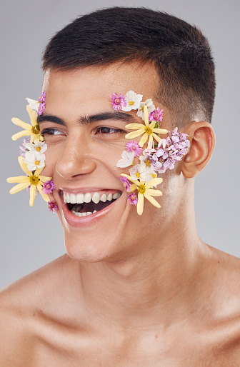 Flowers, man and skincare in studio, cosmetics and creative art on face for spring, beauty and wellness. Closeup, smile and person, makeup and floral decoration for glow skin on gray background