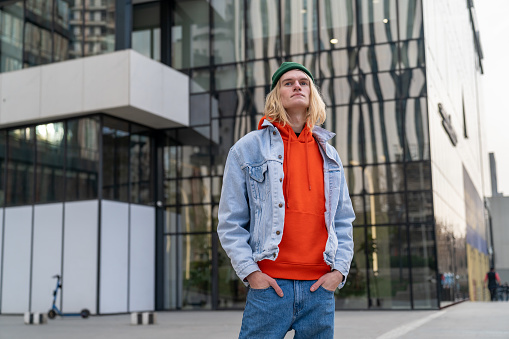 Guy hipster with hands on pockets stands waits somebody on city street near modern glass building. Comfortable urban environment fo living, meeting with friends concept. Blonde man with long hair.