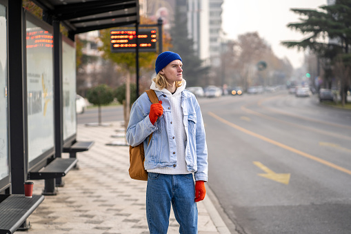 Young guy tourist with backpack waiting at bus stop early in morning. Male passenger use public transport in city. Move without car in big city. Teen waiting bus very long time and looks into distance