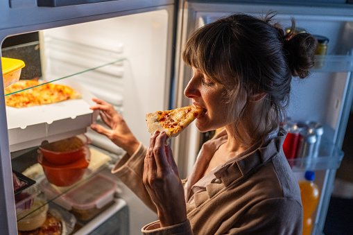 Mature woman eating pizza while standing near open refrigerator at home.