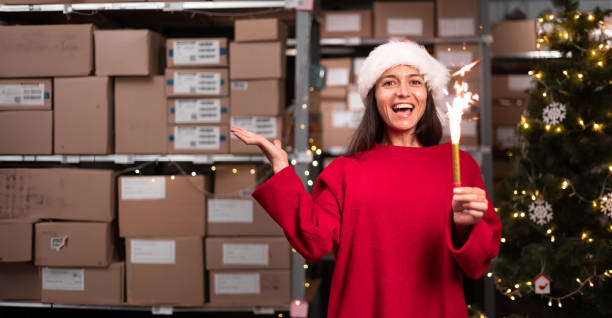 istock Woman holding Merry Christmas fireworks looking at camera laughing standing in workshop warehouse. Fast xmas delivery concept. 1676017708