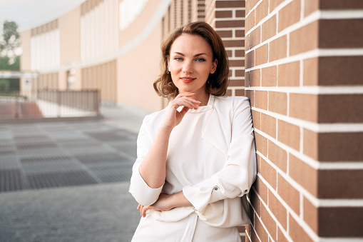 Portrait of smiling confident business woman wearing stylish clothing looking at camera standing near wall. Successful business