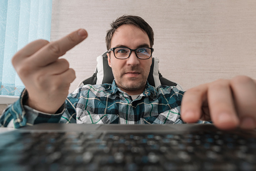 man writes comments while sitting at a laptop and shows his middle finger at a computer screen monitor. insulting and bullying in comments and chats on the Internet
