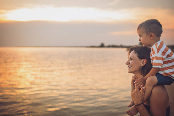 Woman carrying her little son on shoulders on the beach by the sea, they are on vacation.