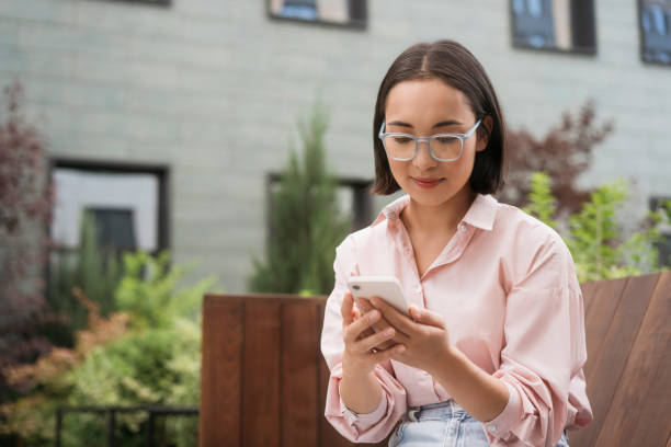 istock Asian woman wearing eyeglasses using mobile phone, reading text message sitting on bench in park 1676160047