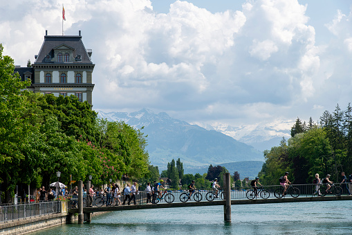 Thun, Switzerland-May 28, 2023; Cyclists over bridge in the Aare river in historic downtown with Lake Thun and snowcapped mountains in the background