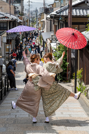 Kyoto, Japan-April 14, 2023; Close up of two girls in traditional kimono making a pose with umbrella’s raised in the air in the street of historic Higashiyama district