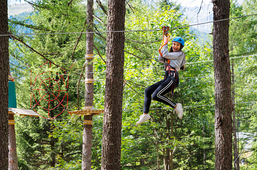 Side view of excited young female with opened mouth looking down while having fun on high ropes with belay cables protective helmet in forest adventure park in daylight