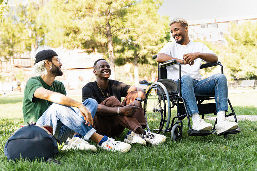 Disabled man on wheelchair together with young friends having fun sitting on park grass, talking and laughing