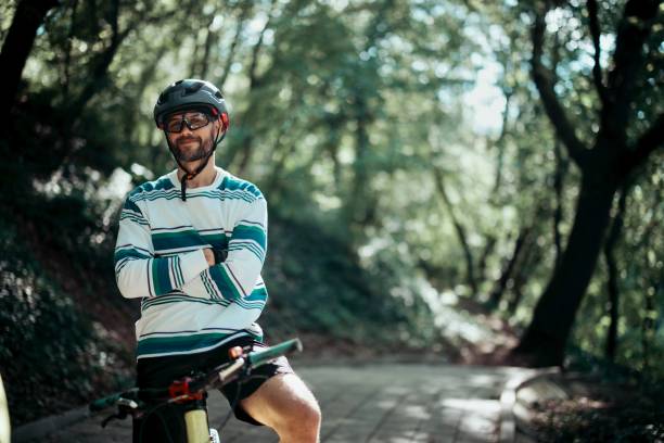 A caucasian man in a black t-shirt and shorts is staying with a black bike in a forest in the orange sunset light