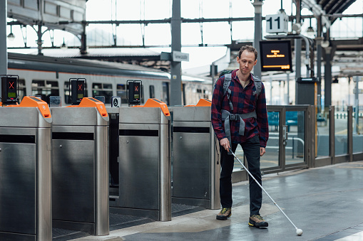 The image shows a partially sighted man leaving a railway station using his cane. In the background are the ticket gates and a stationary train.