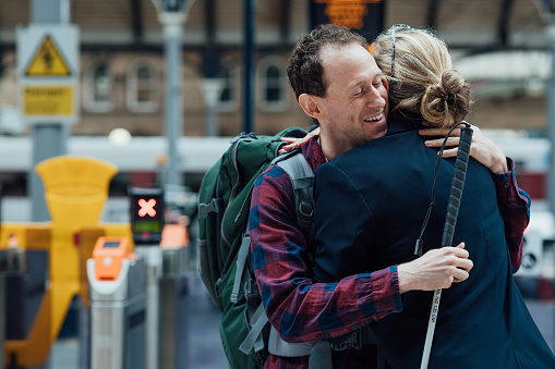 Two friends are hugging at a railway station in northeast England. One is visually impaired and is holding his white cane in his hand. He is wearing a rucksack. In the background, one can see a train. The station is an open canopy from the time of steam trains.