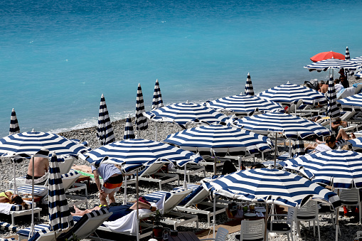 Nice, France - April 26, 2023: A Mediterranean beach with blue and white umbrellas. There are beachgoers here who enjoy the seaside charms in the shade of umbrellas