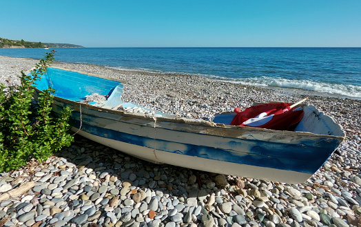 An abandoned and broken boat and the same chair lies on the shore of the Aegean Sea, surrounded by bushes and trees with a house in the distance in Greece