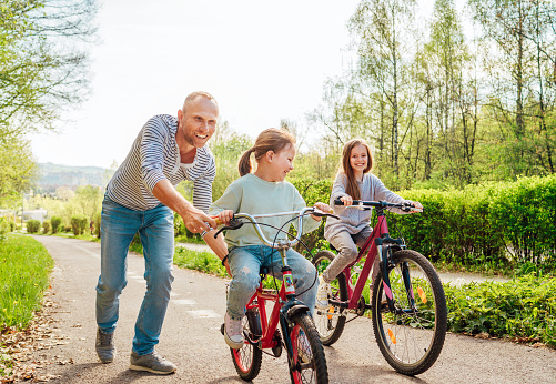 Smiling father with two daughters during outdoor walk. He teaching younger girl to ride a bicycle. They enjoy togetherness in the summer city park. Happy childhood concept image.